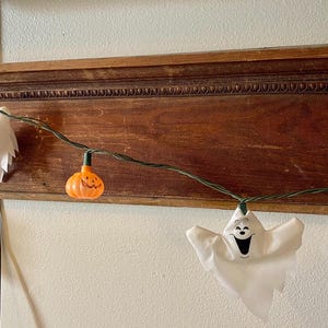 May include: A Halloween-themed string light set featuring white ghost and orange pumpkin-shaped lights. The lights are strung on a green wire and are displayed against a wooden shelf and white wall.