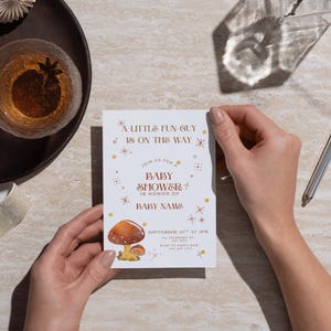 May include: A baby shower invitation with the text "A Little Fun Guy Is On The Way." The invitation features a mushroom illustration and is held by a person. A pen, a glass of liquid, and decorative items are also visible.