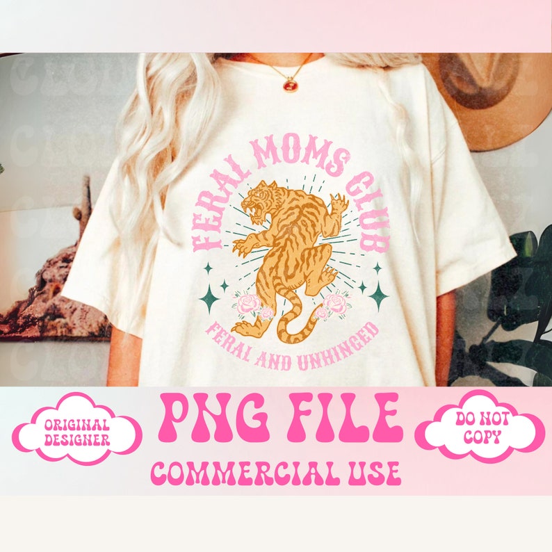 Op de afbeelding: Een wit T-shirt met een roze en groen grafisch ontwerp met een tijger en de tekst "Feral Moms Club" en "Feral and Unhinged".