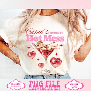 以下が含まれることがあります： ピンクと赤のグラフィックデザインが施された白いTシャツで、「Cupid's Favorite Hot Mess」と書かれています。グラフィックには、翼のある漫画のキューピッド、赤いカクテルグラス、ピンクのディスコボールハートが描かれています。