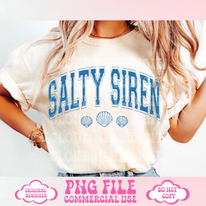May include: Cream-colored t-shirt with the text "SALTY SIREN" in blue, arched across the chest. Below the text are three blue seashell graphics. The shirt has rolled-up sleeves and is worn by a person.