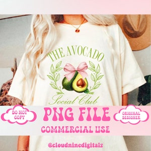 以下が含まれることがあります： クリーム色のTシャツ。緑とピンクのアボカドのデザインと「The Avocado Social Club」の文字がプリントされています。グラフィックにはピンクのリボンと緑の葉が含まれています。シャツには「PNG FILE COMMERCIAL USE」の文字も入っています。