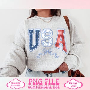 May include: A gray sweatshirt with the word "USA" printed on the front. The letters are decorated with red, white, and blue patterned fabric. A blue ribbon with a bow is below the letters.