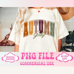 Op de afbeelding: Een wit T-shirt met de tekst "AUTUMN Girly" in een kleurrijk, gelaagd lettertype. De tekst is in een varsity-stijl lettertype met een bruin, oranje, rood en groen kleurenschema. Het shirt wordt gemodelleerd door een persoon met lang blond haar.