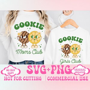 May include: Two white t-shirts with a graphic design of a cookie and a lemon slice with faces. The text on the shirts reads "Cookie Mom's Club" and "Cookie Girls Club".
