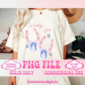 Puede incluir: Un diseño digital que presenta dos herraduras rosas con lazos azules y mariposas. El texto "Lucky You" está escrito en cursiva sobre las herraduras. El diseño está en una camiseta de color crema.
