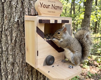 May include: A light-colored wooden squirrel feeder shaped like a food truck with a sign that reads "OPEN 24 HRS Your Name FOOD TRUCK". The feeder features a dark wood accent, a small wheel, and a platform where a gray squirrel eats seeds.