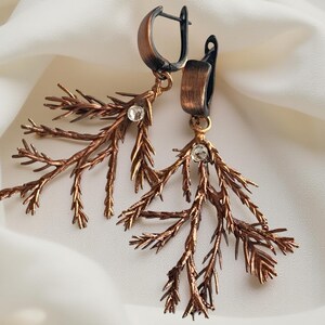 May include: A pair of copper-colored earrings with a botanical design. Each earring features a branch-like structure with small, clear crystals and a hinged clasp. The earrings are set against a white, draped fabric.