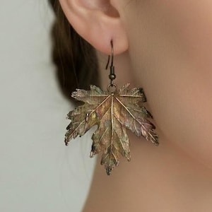 May include: A dangling earring shaped like a maple leaf with a metallic bronze and copper finish. The leaf features intricate, realistic vein detailing across its surface and is suspended from a thin, dark metal hook wire.