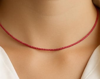 May include: A delicate, red beaded necklace is worn against a white collared shirt. The necklace is a thin strand of small, evenly spaced beads. The shirt has a logo on the lower right side. The necklace is a simple, elegant accessory.
