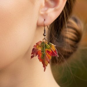 May include: A drop earring featuring a realistic maple leaf pendant with a gradient of autumn colors including red, orange, yellow, and green. The leaf is suspended from a dark metal hook, showcasing intricate vein details and a textured surface.