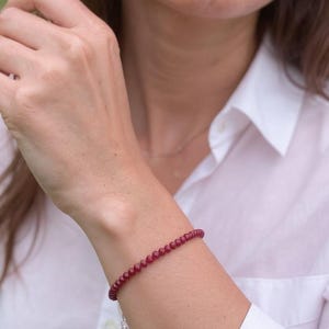 May include: A burgundy beaded bracelet with a silver chain and clasp. The bracelet is worn on a wrist, and the beads appear to be small and round. The background is blurred, suggesting an outdoor setting with greenery.