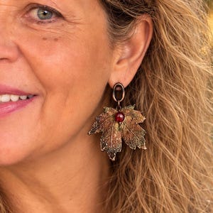 May include: A dangling earring featuring a textured, metallic maple leaf in shades of bronze, copper, and dark green. The leaf is accented with a small, round, deep red bead at the top, suspended from a simple dark metal oval hoop attachment.