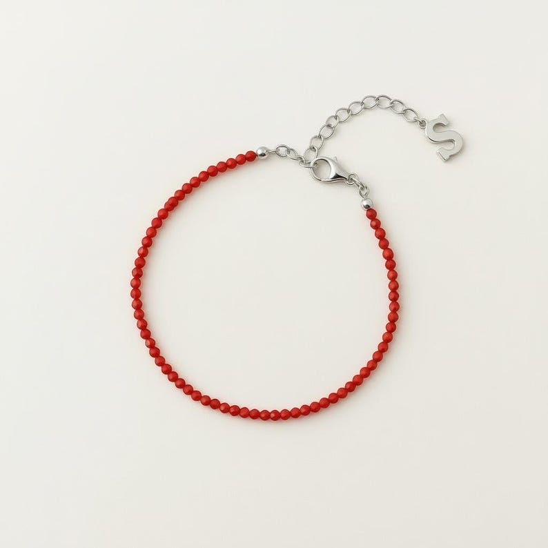 K&ouml;nnte beinhalten: Ein zartes Armband mit kleinen, runden, leuchtend roten Perlen auf einem d&uuml;nnen Draht. Das Armband verf&uuml;gt &uuml;ber einen silberfarbenen Karabinerverschluss, eine kurze Verl&auml;ngerungskette und einen kleinen Anh&auml;nger in Form des Buchstabens S.