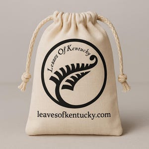 K&ouml;nnte beinhalten: Ein beigefarbener Baumwollbeutel mit Kordelzug, der ein schwarzes kreisf&ouml;rmiges Logo mit einer stilisierten Farnblattgrafik und dem Text Leaves Of Kentucky zeigt. Unter dem Logo ist der Text leavesofkentucky.com in einer schwarzen Serifenschrift.