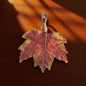 May include: A maple leaf pendant featuring a textured surface with a gradient of deep red, orange, and yellow hues. The leaf has a metallic bail attached to the top stem, showcasing intricate vein details across the entire surface of the leaf.