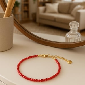 K&ouml;nnte beinhalten: Ein zartes Armband mit kleinen, facettierten roten Perlen auf einer d&uuml;nnen Schnur. Das Schmuckst&uuml;ck verf&uuml;gt &uuml;ber einen goldfarbenen Karabinerverschluss, eine verstellbare Verl&auml;ngerungskette und einen kleinen goldfarbenen Muschelanh&auml;nger.