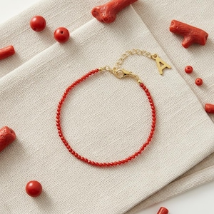 May include: A delicate bracelet composed of small, round red beads strung on a gold-toned chain. The bracelet features a gold-toned lobster clasp and a dangling charm in the shape of the capital letter A. Several loose red beads and coral pieces.