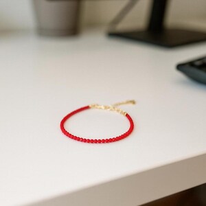 K&ouml;nnte beinhalten: Ein zartes Armband bestehend aus einer einzelnen Reihe kleiner, leuchtend roter Perlen. Das Armband verf&uuml;gt &uuml;ber eine goldfarbene Metallkettenverl&auml;ngerung mit einem Verschluss, die eine individuelle Anpassung an das Handgelenk erm&ouml;glicht.