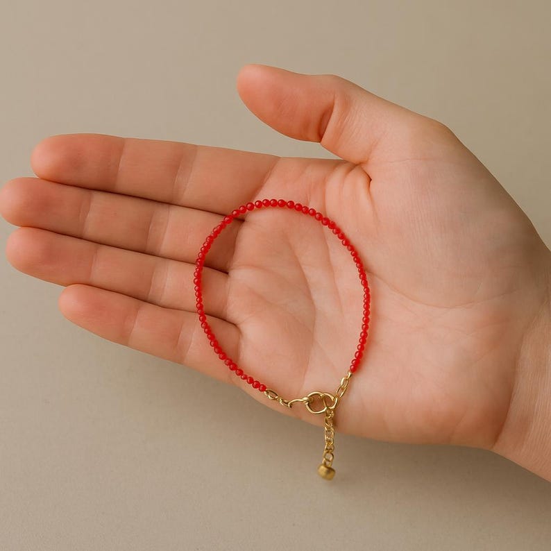 K&ouml;nnte beinhalten: Ein zartes Armband mit einer durchgehenden Reihe kleiner, leuchtend roter Perlen. Das Armband verf&uuml;gt &uuml;ber eine goldfarbene Metallkettenverl&auml;ngerung mit einem Karabinerverschluss und einem kleinen runden Anh&auml;nger am Ende zur Gr&ouml;&szlig;enanpassung.