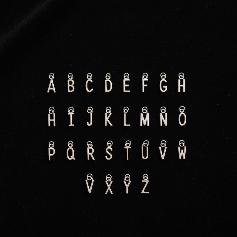 K&ouml;nnte beinhalten: Eine Sammlung von sechsundzwanzig einzelnen silberfarbenen Metall-Buchstabenanh&auml;ngern, die jeweils einen Gro&szlig;buchstaben von A bis Z zeigen. Jeder Buchstabenanh&auml;nger ist mit einem kleinen runden Biegering an der Oberseite versehen.