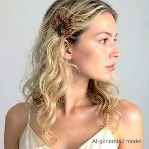 May include: A hair clip shaped like a maple leaf with a metallic bronze and dark brown finish. The leaf features intricate veining and a textured surface, secured in wavy blonde hair. The accessory is positioned on the side of the head.