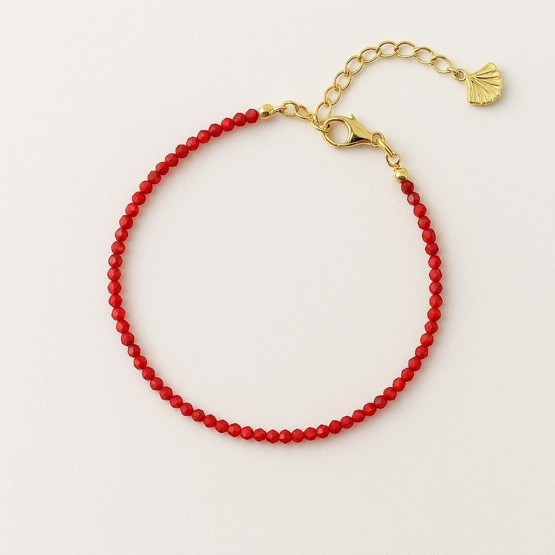 K&ouml;nnte beinhalten: Ein zartes Armband mit kleinen, facettierten roten Perlen an einer goldfarbenen Kette. Das Schmuckst&uuml;ck verf&uuml;gt &uuml;ber einen goldfarbenen Karabinerverschluss, eine verstellbare Verl&auml;ngerungskette und einen kleinen f&auml;cherf&ouml;rmigen Anh&auml;nger.