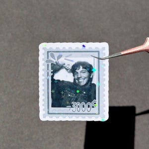 May include: A rectangular holographic sticker shaped like a postage stamp featuring a black and white portrait of a smiling person with one arm raised. The sticker includes the text -3000 and has a scalloped edge with a shimmering star pattern.