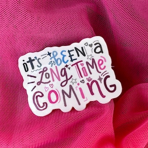 May include: White sticker with the text "it's been a long time coming" in black, pink, and blue lettering with decorative stars and hearts.