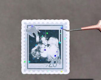 May include: A rectangular sticker shaped like a postage stamp with a scalloped edge. The centre features a black and white photograph of three people, a small tooth icon, and the text 20$. The sticker has a holographic finish with star accents.