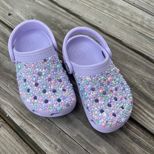 May include: A pair of lavender clogs decorated with iridescent rhinestones and beads. The clogs have a strap on the back and are made of a soft, comfortable material.