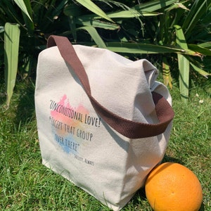 May include: A white canvas tote bag with brown straps. The bag has a rainbow watercolor design and the text "Unconditional Love! *Except that group over there* Society. Always". The bag is sitting on green grass next to an orange.