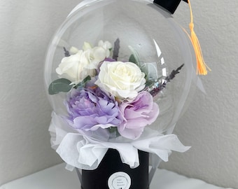 Peut inclure: Un bouquet de ballons transparent avec un bonnet de graduation sur le dessus. Le bouquet contient des fleurs blanches et violettes et se trouve dans une boîte noire avec un ruban blanc. Le texte sur la boîte indique "Happy Balloon Blooms".