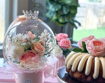 Peut inclure: Une sphère en verre transparente surmontée d'une couronne rose, contenant un bouquet de fleurs roses et blanches. La sphère est posée sur une boîte rose avec une étiquette blanche. Un gâteau blanc avec un glaçage blanc est sur un support à gâteau blanc.
