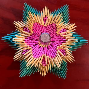 Può includere: Un colorato fiore origami con un elemento centrale in vetro. Il fiore presenta un design a stella con triangoli di carta rosa, gialli, verdi e blu. Un oggetto decorativo.