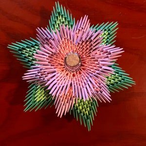 May include: A colorful, origami-style flower with a central glass element. The flower features layers of folded paper in shades of pink, green, and orange, creating a starburst design. The object is placed on a brown surface.