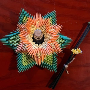Może przedstawiać: Kolorowy kwiat origami z zielonymi, pomarańczowymi i żółtymi płatkami. Kwiat jest wykonany z papieru i ma szklaną butelkę w środku. Kwiat leży na drewnianej powierzchni. Dwie czarne pałeczki z małym kwiatem origami na końcu znajdują się w tle.