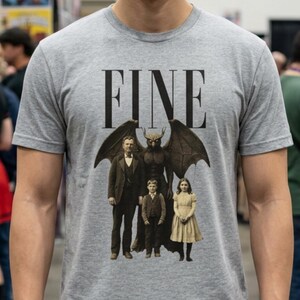 May include: A gray t-shirt with the word "FINE" printed in black above a graphic of a family with a winged demon. The shirt is short-sleeved and worn by a person in a convention setting.