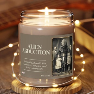 May include: A brown candle with a white label that reads "Alien Abduction". The label features a black and white photograph of a group of people with a large alien figure in the centre. The candle is on a wooden coaster and surrounded by white fairy lights.