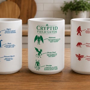 Puede incluir: Tres tazas de cerámica blanca con asas, cada una con ilustraciones y texto de críptidos. Una taza tiene gráficos azules de Bigfoot, Nessie y el Monstruo de Flatwoods. Otra tiene gráficos verdes y el texto "Cryptid Field Guide".
