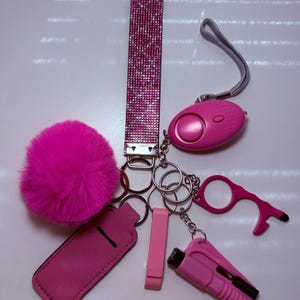 May include: A pink keychain with a variety of safety and utility tools, including a personal alarm, a door opener, a lighter, and a pom-pom. The keychain also features a pink sparkly strap.