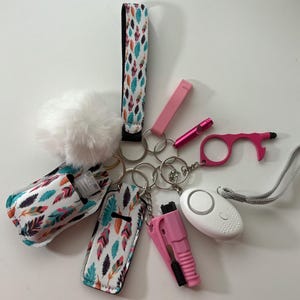 Può includere: Una collezione di articoli per la sicurezza personale, tra cui un portachiavi con un pompon bianco, un cinturino con un motivo a piume e vari accessori rosa e bianchi. Gli articoli includono un allarme personale, un apriporta e un taglia cinture.
