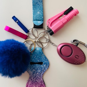 Puede incluir: Un llavero de purpurina azul y rosa con un pompón azul, un silbato, un corta-cinturones rosa y una alarma personal rosa.