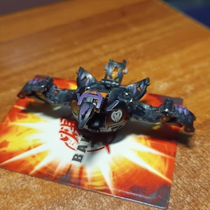 Peut inclure: Un jouet Bakugan noir et violet avec des accents orange. Le jouet est en position fermée et a un symbole argenté sur le devant. Le jouet est sur une carte avec le mot "Bakugan" imprimé dessus.