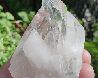 May include: A large, clear quartz crystal cluster with multiple points. The crystal is translucent and has a milky white color. The cluster is sitting on a hand.