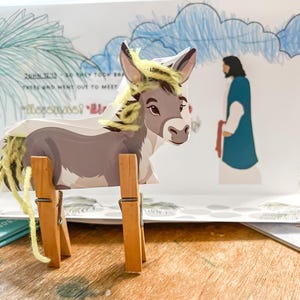 Peut inclure: Un âne gris avec des cheveux en laine jaune et un support en bois brun. L'âne est debout devant une carte blanche avec un ciel bleu et une personne en robe bleue. La carte porte le texte "Jean 12:13 - Alors ils prirent des branches de palmiers et allèrent à sa rencontre."