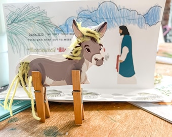 May include: A gray donkey with yellow yarn hair and a brown wooden stand. The donkey is standing in front of a white card with a blue sky and a person in a blue robe. The card has the text "John 12:13 - So they took branches of palm trees and went out to meet him."