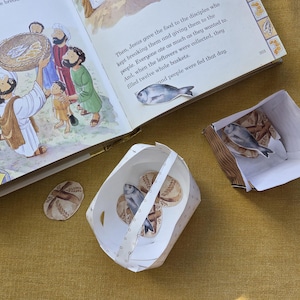 Könnte beinhalten: Ein offenes Buch mit einer Illustration von Menschen und Essen. Neben dem Buch befinden sich Papierschnitte von Brot und Fisch, zusammen mit einem Papierkorb und einem Papierbehälter. Die Szene spielt sich auf einer gelben Oberfläche ab.