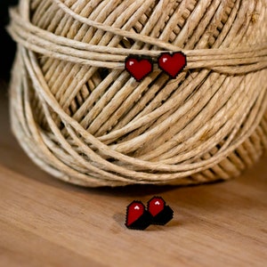 May include: Red pixel heart earrings on a spool of twine. The earrings are small, with a black border and a red heart design. The twine is a natural beige color, and the background is a wooden surface.