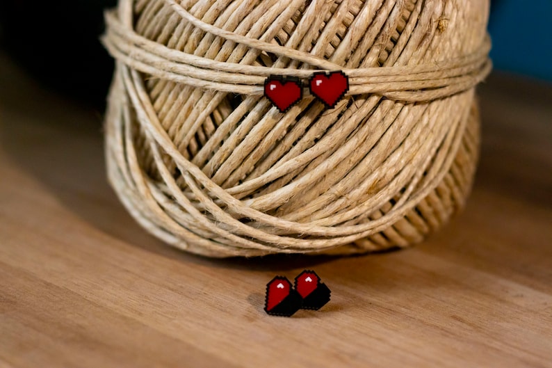 May include: Red pixel heart earrings on a spool of twine. The earrings are small, with a black border and a red heart design. The twine is a natural beige color, and the background is a wooden surface.